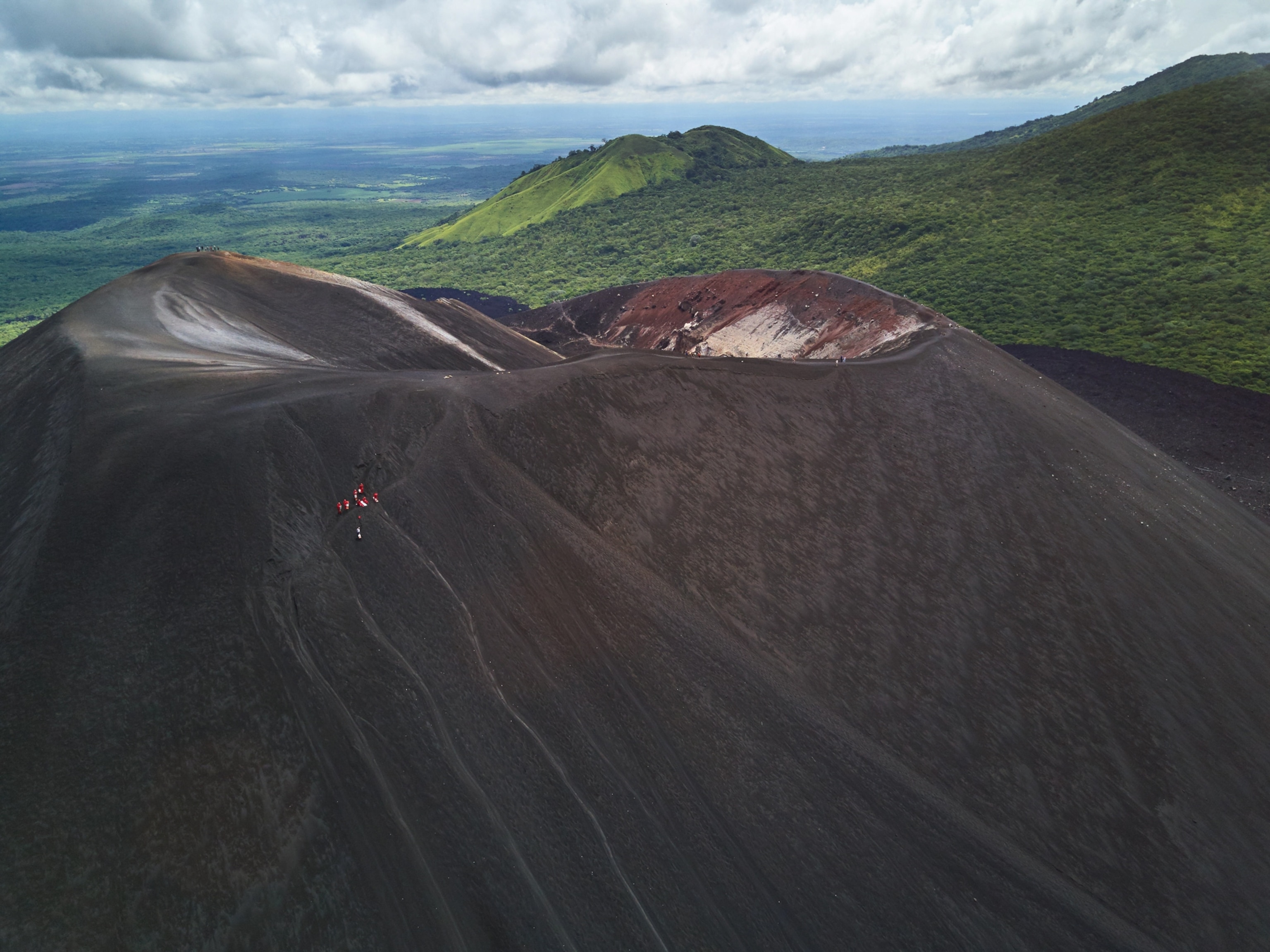 An elevated view of an ash-covered volcano with green hills in the background. Near the top of the volcano, a group of people appear minuscule as they gather to ride boards down the volcano.