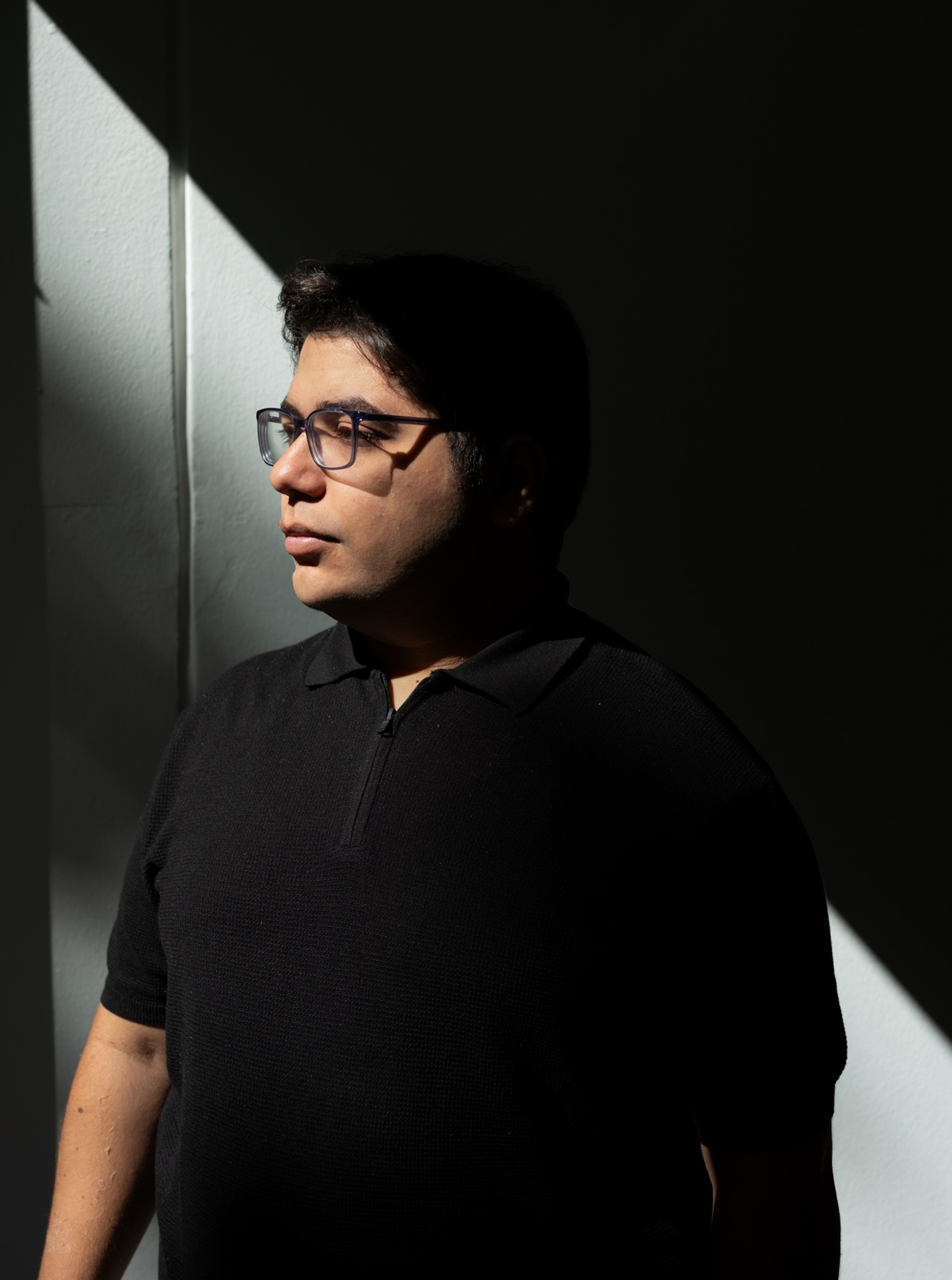 A man with glasses and a dark shirt stands amidst a shadowed wall for a profile shot.