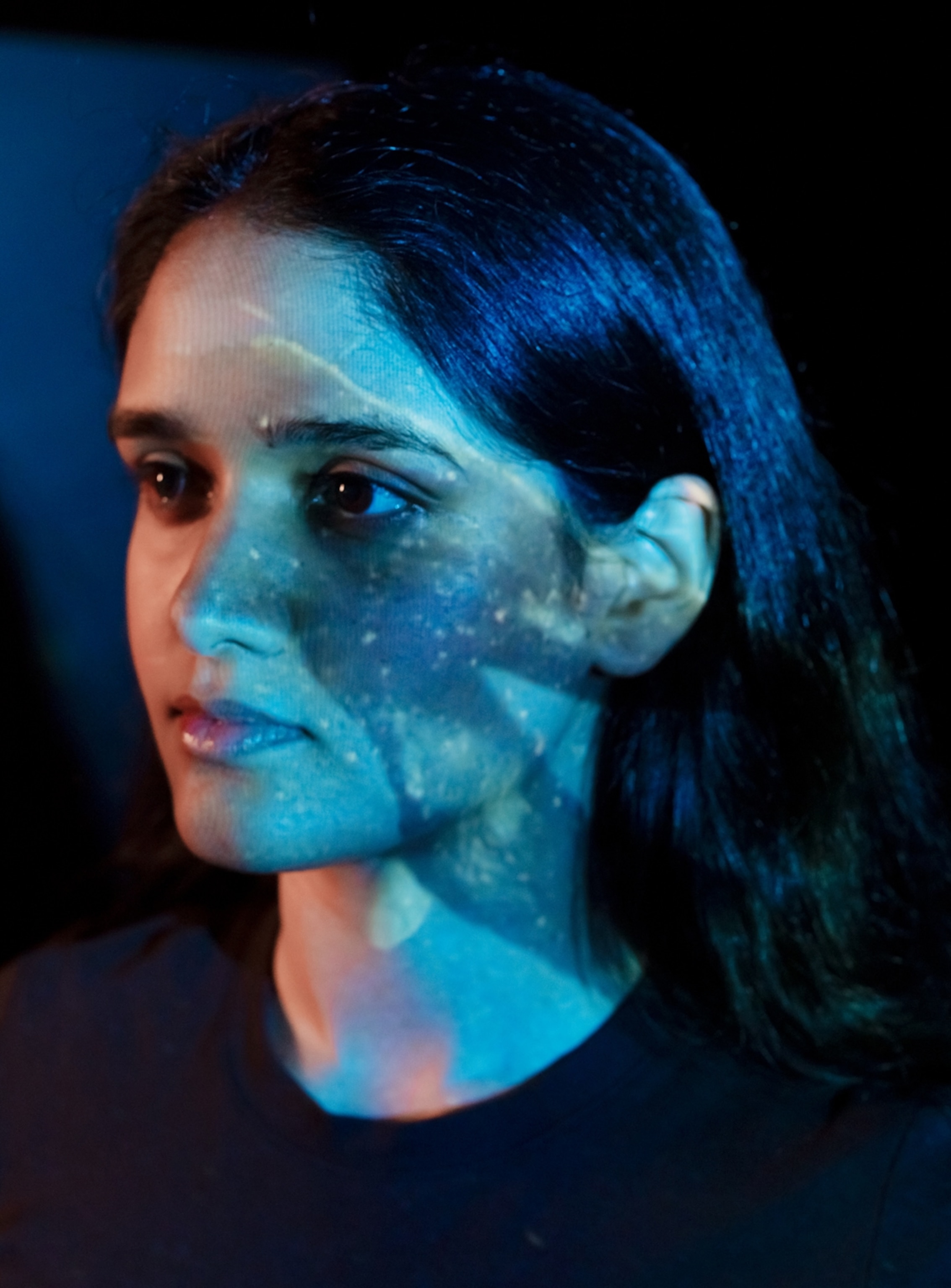 A profile of a woman with dark features and a dark shirt. She has water-like reflections across her face.