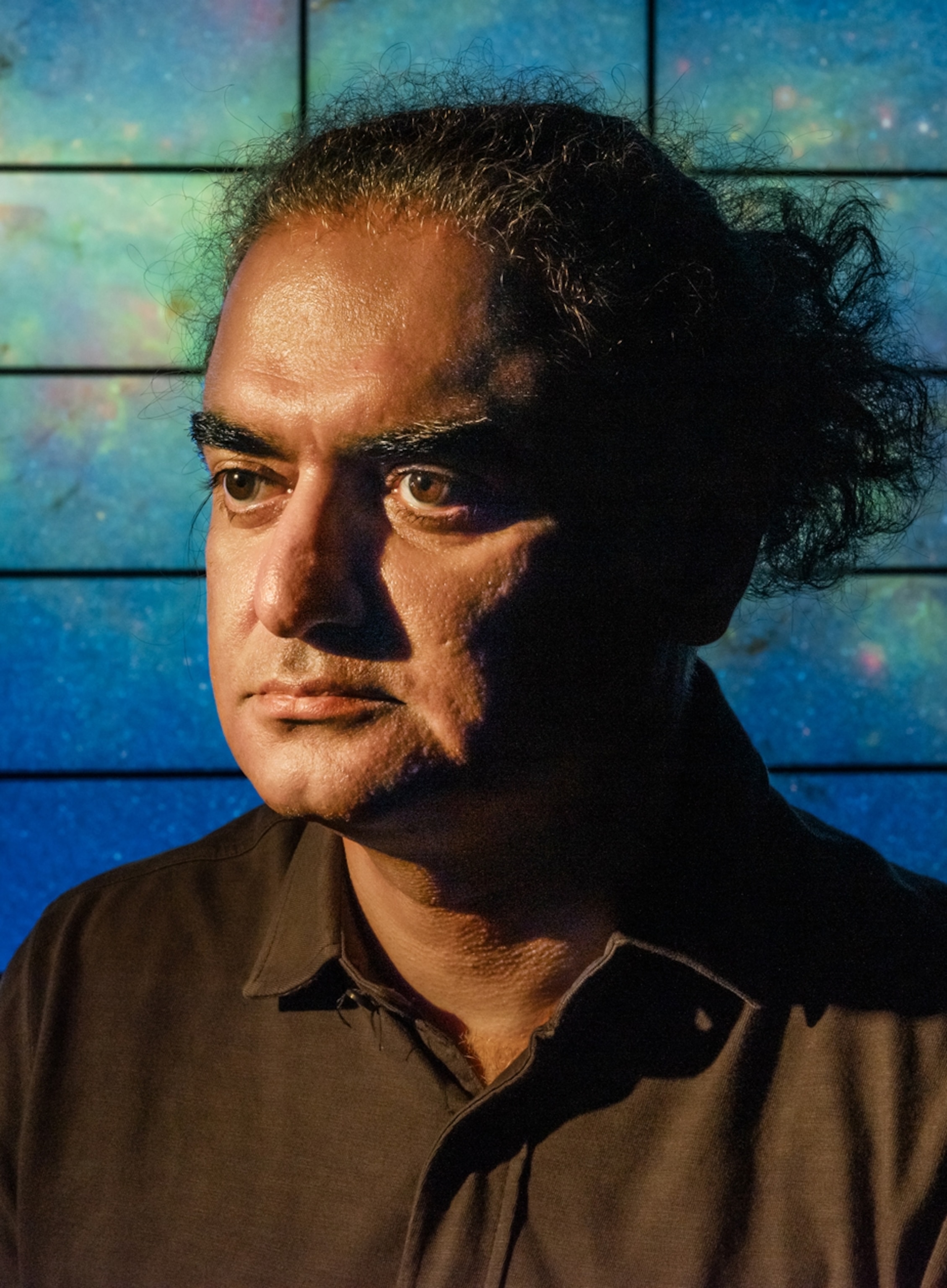 A close-up portrait of a man. He has a brown shirt and black hair tied back. There is a galaxy-like background behind him.