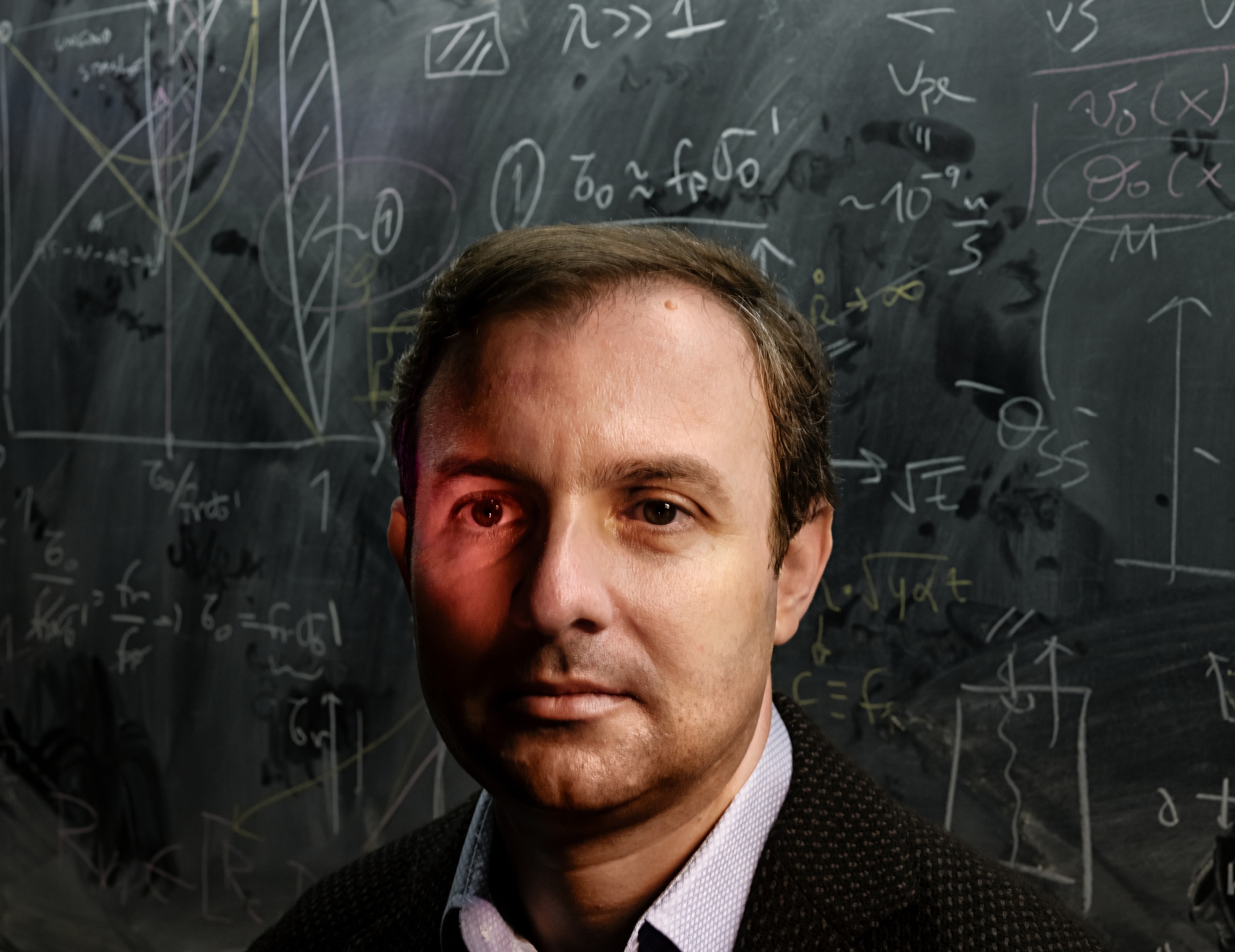 A portrait of a man with short brown hair. There is a red light shining on his right eye and there is a chalk board with equations written on it behind him.