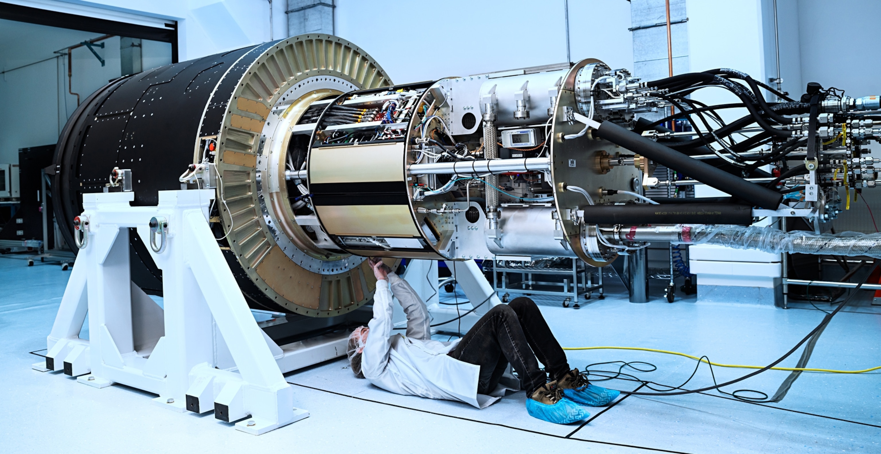 A man in a white coat and blue shoe covers is seen laying on his back underneath a large piece of equipment. This equipment is cylindrical in shape, and gold and black. It is the largest camera on Earth.