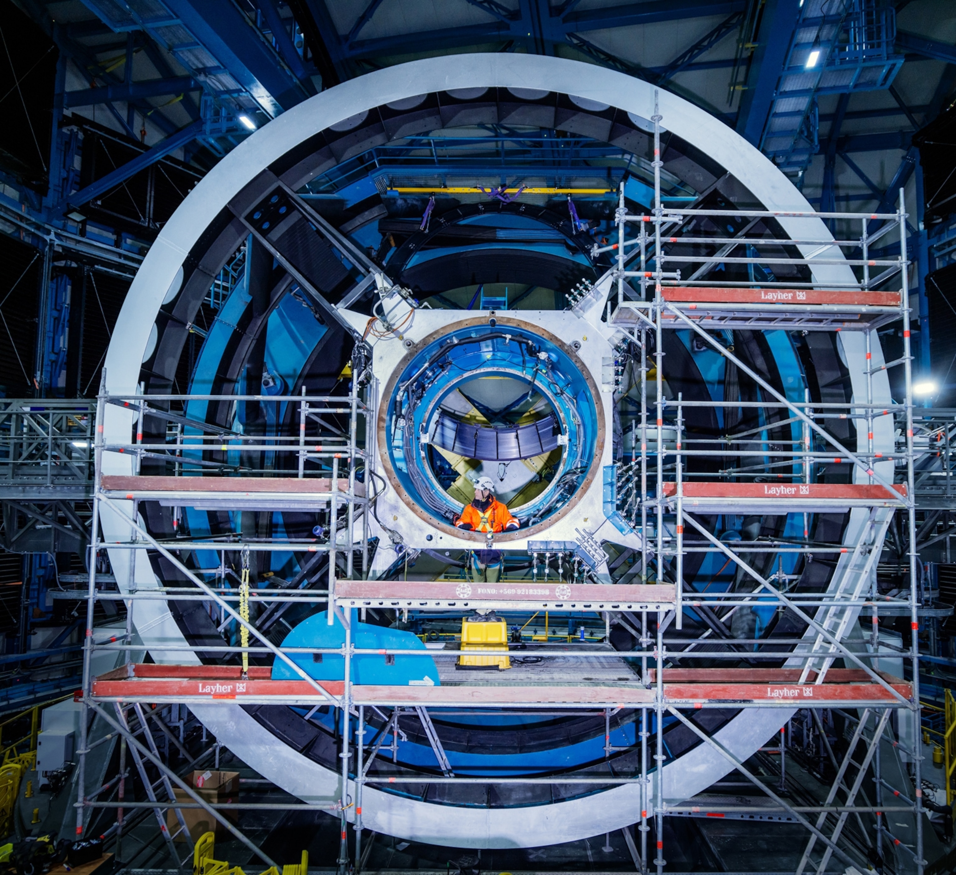 A man in an orange and yellow construction uniform is seen inside a large circular infrastructure. This is the mount for an extremely large mirror which will be part of the Vera Rubin telescope.