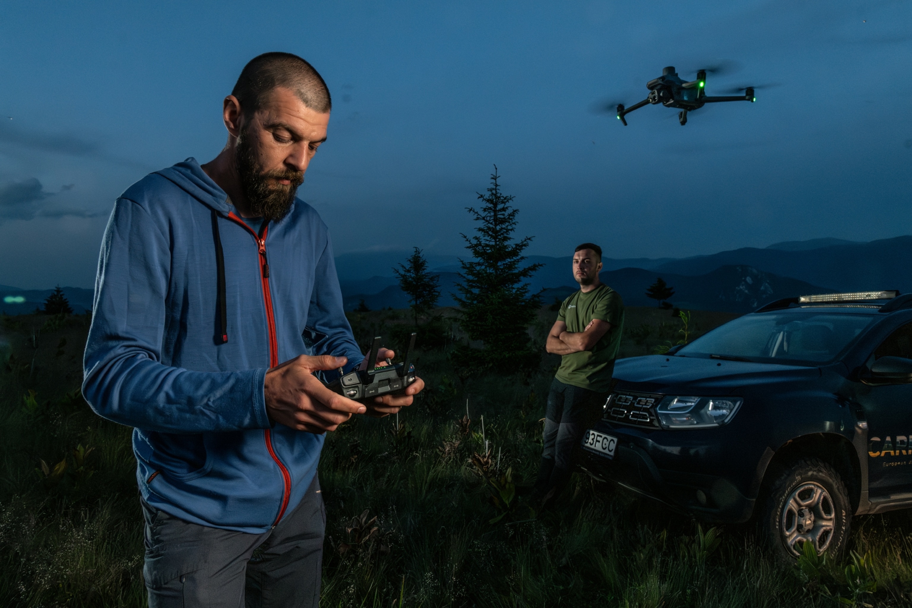 Sebastian is controlling a drone flying next to him on the right. Another man is watching, while leaning on the hood of a car behind.