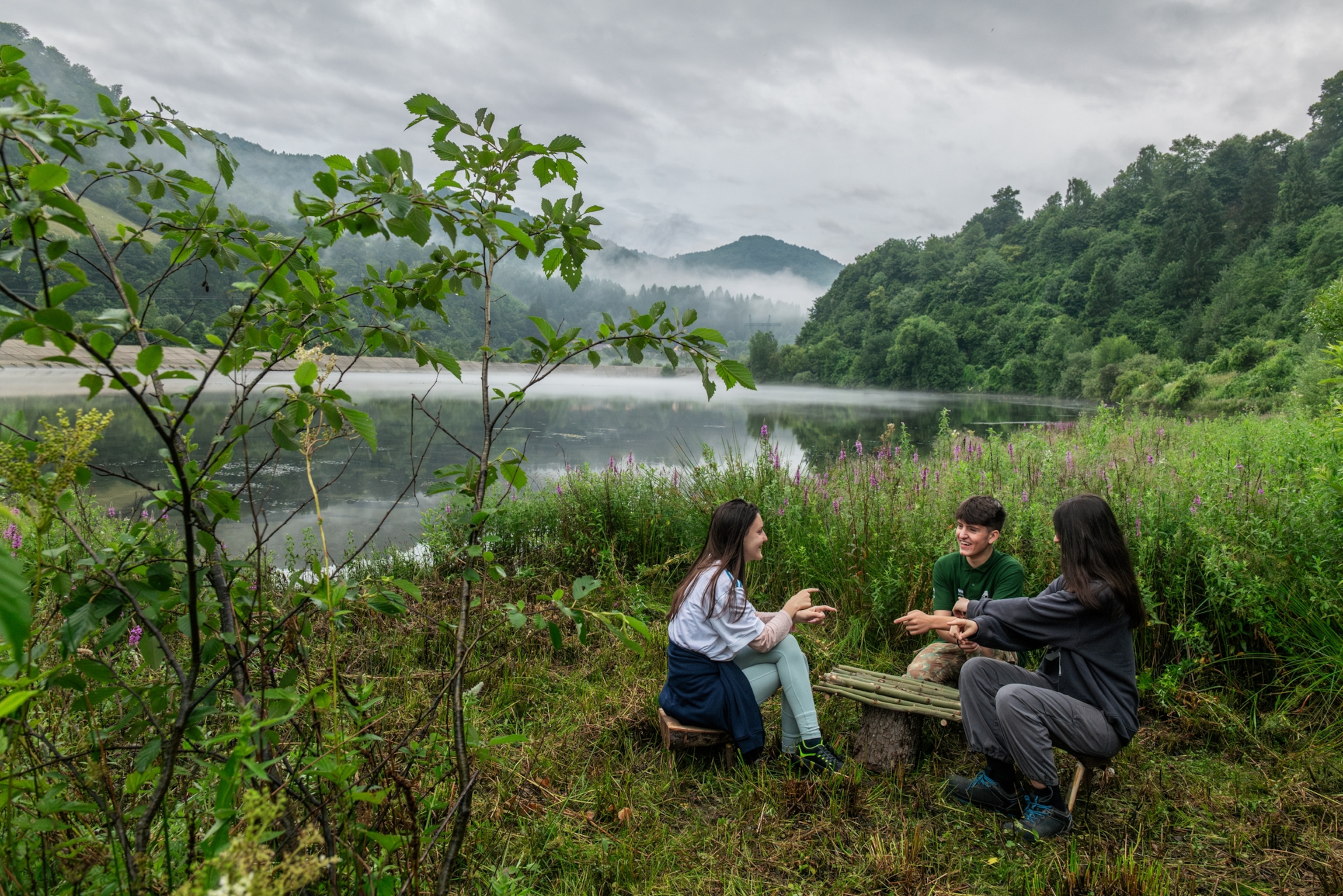 three people sitting at a self-made table with chairs next to the water and surrounded by lush forest