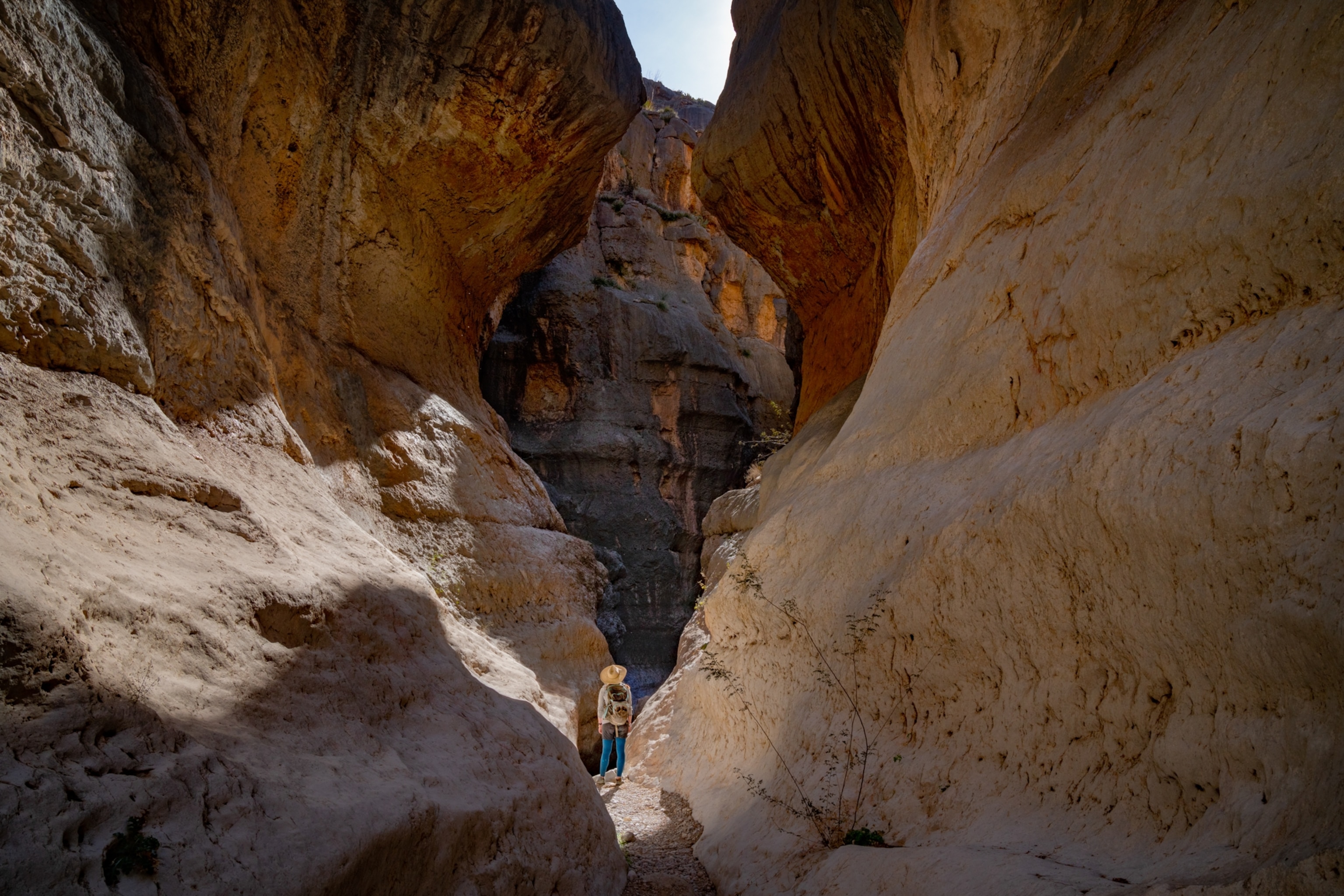 A lone hiker appears tiny as she looks up and walks between the walls of a stone gorge.