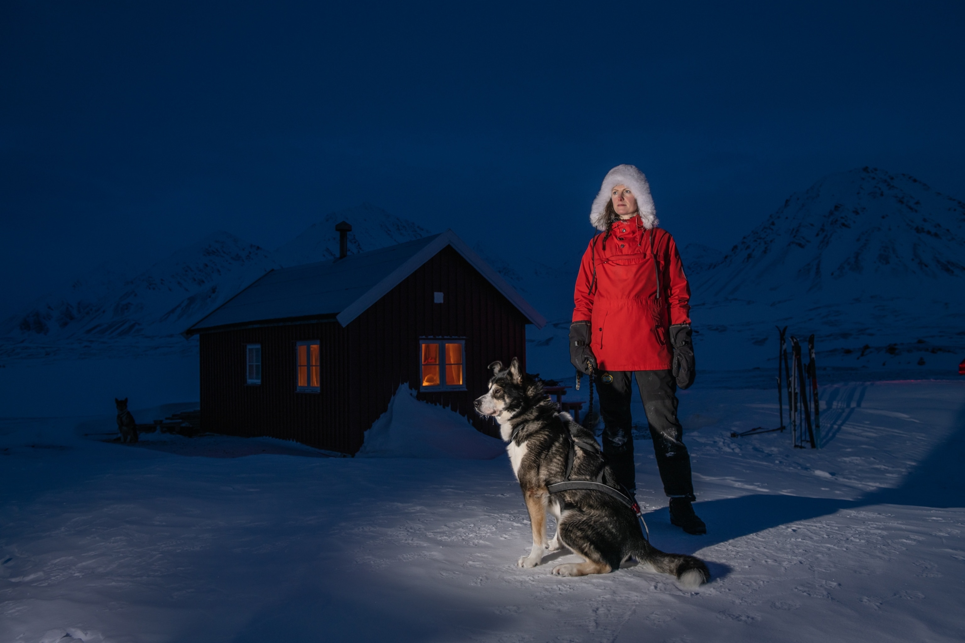 Portrait of Kjerstad in a red parka holding her dog in the snow. She is standing in front of a warmly lit hut with skiis outside.