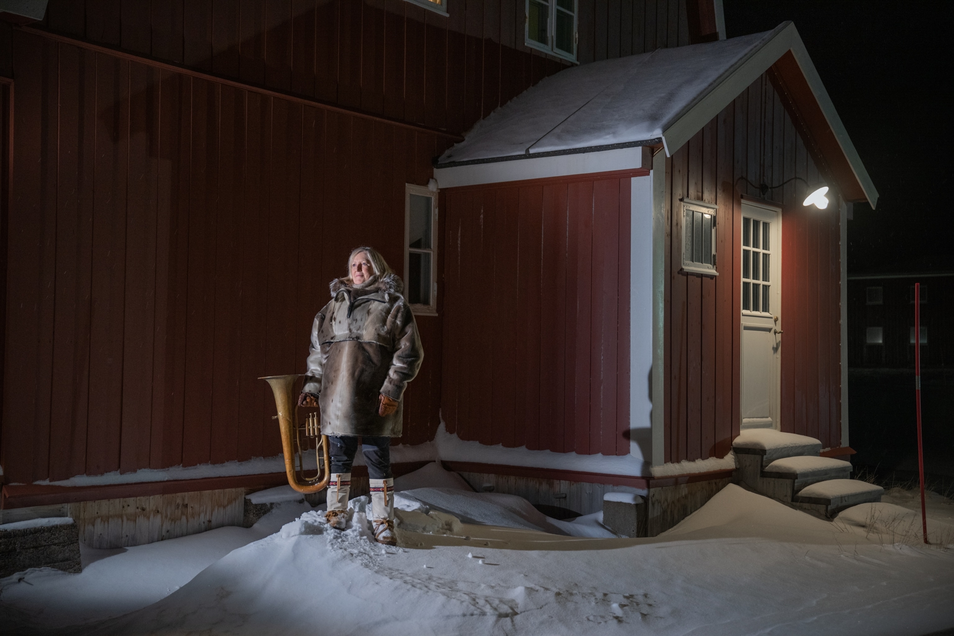 Portrait of Haugvik warmly dressed, outside a snowy, red wood house holding a baritone horn.