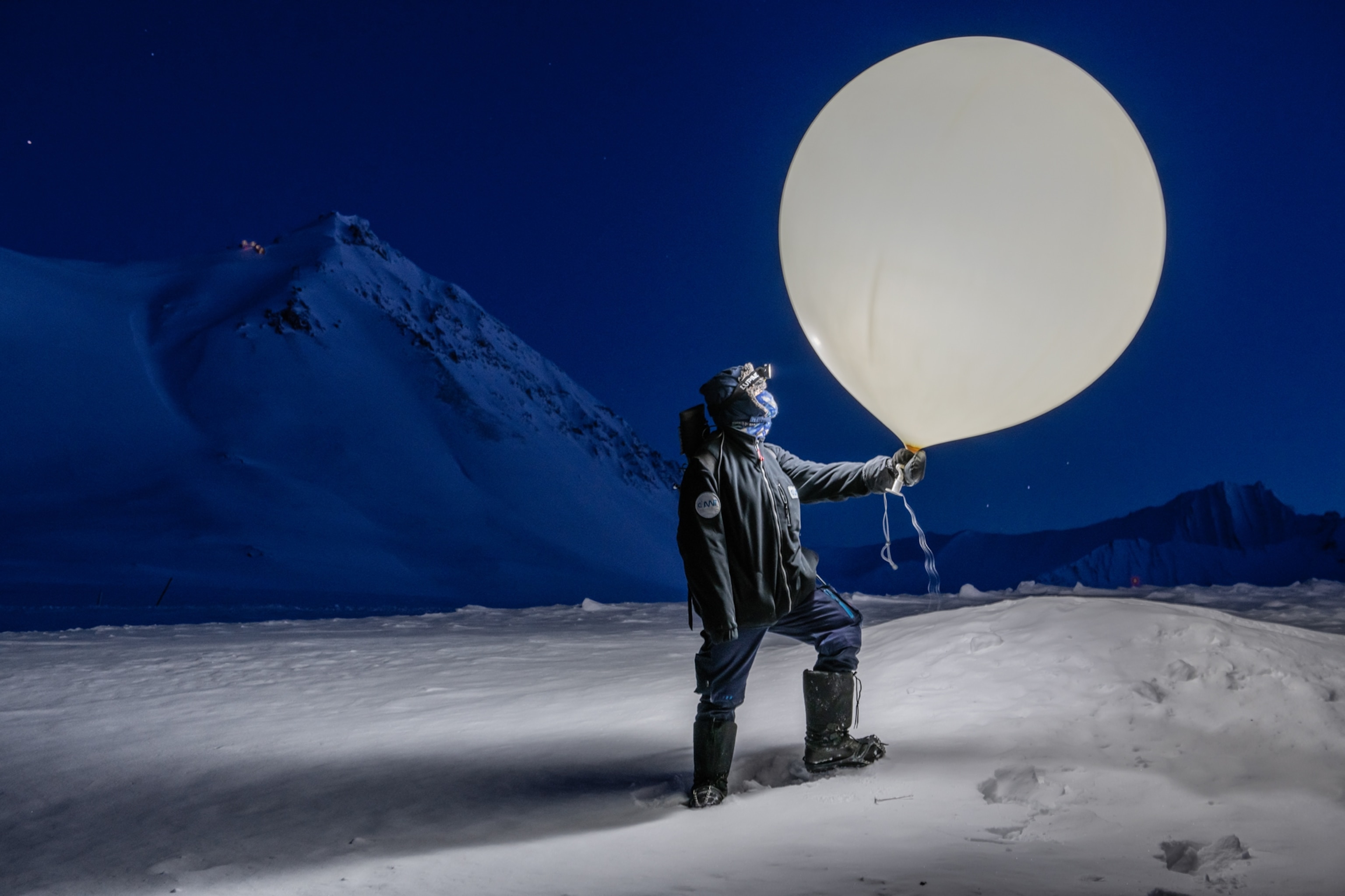 Portrait of Haupt standing outside in snow holding a large white weather balloon. Haupt is dressed warmly and is wearing a headlamp